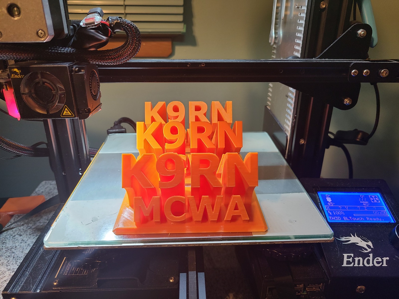 3D printer making orange plastic models with "K9RN" and "MCWA" on a printing bed, Ender label visible.