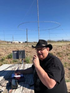 Man with black hat and glasses holding a microphone at an outdoor table with radio equipment.